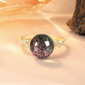 Könnte beinhalten: Ein goldener Ring mit einem großen, runden Edelstein mit grünen und roten Farbtönen. Der Ring ist mit kleinen, klaren Akzentsteinen besetzt. Der Ring ist vor einem weichen, warmen Hintergrund platziert.