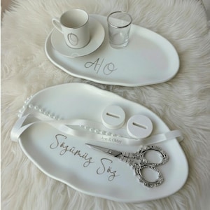 May include: Two white oval serving trays with silver accents. One tray holds a small teacup and saucer, and a shot glass. The other tray has scissors, two white ring holders, and a ribbon. The trays have script writing.