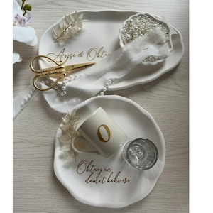 May include: Two white, irregularly shaped serving trays with gold text and decorative items. One tray has gold scissors, pearls, and a small dish of pearls. The other has a mug with a gold "0" and a small glass.