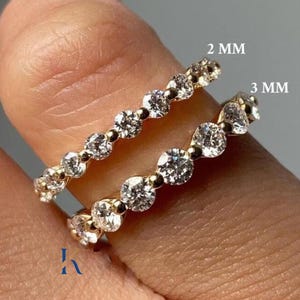 Könnte beinhalten: Zwei goldene Ringe mit klaren Edelsteinen. Ein Ring ist mit "2 mm" und der andere mit "3 mm" beschriftet. Die Ringe befinden sich an einem Finger. Die Ringe sind wahrscheinlich für eine Hochzeit oder einen Verlobung.