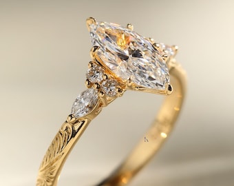 May include: A gold ring featuring a large marquise-cut center stone, flanked by two smaller round stones and a marquise-cut stone on each side. The band is crafted from yellow gold and displays an intricate, engraved leaf-like pattern along its surface.