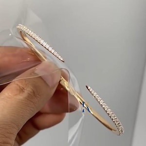 May include: A gold-toned open cuff bracelet featuring two tapered ends, each encrusted with a row of small, clear, sparkling stones. The bracelet has a smooth, polished finish and a flexible design that allows for an adjustable fit on the wrist.