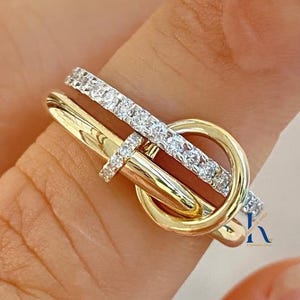 Puede incluir: Un anillo bicolor que presenta una banda de oro amarillo pulido con forma de hebilla y una banda de tono plateado incrustada con pequeñas piedras transparentes. El diseño incorpora un elemento circular donde ambas bandas se cruzan.