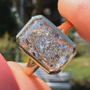 Op de afbeelding: Een gouden ring met een grote, rechthoekig geslepen diamant. De heldere diamant schittert. De achtergrond is wazig en groen.