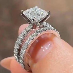 May include: A silver engagement ring set with a large, square-cut diamond. The band is encrusted with smaller diamonds, creating a sparkling effect. The ring is held up, showcasing its intricate design and brilliance.