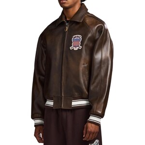 May include: A dark brown leather bomber jacket featuring a front zip fastening, a pointed collar, and ribbed cuffs and hem with white stripes. An embroidered patch on the chest displays the text AVIREX above a stylised American flag design.