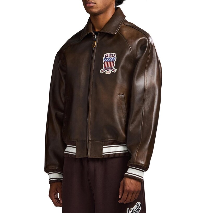 May include: A dark brown leather bomber jacket featuring a front zip fastening, a pointed collar, and ribbed cuffs and hem with white stripes. An embroidered patch on the chest displays the text AVIREX above a stylised American flag design.
