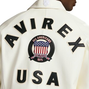 Op de afbeelding: Een gebroken witte jas met zwarte leren letters die AVIREX en USA op de achterkant vormen. Een ronde geborduurde patch in het midden toont een schild met sterren en strepen, en de tekst ALL AMERICANS, ESTABLISHED en 1975.