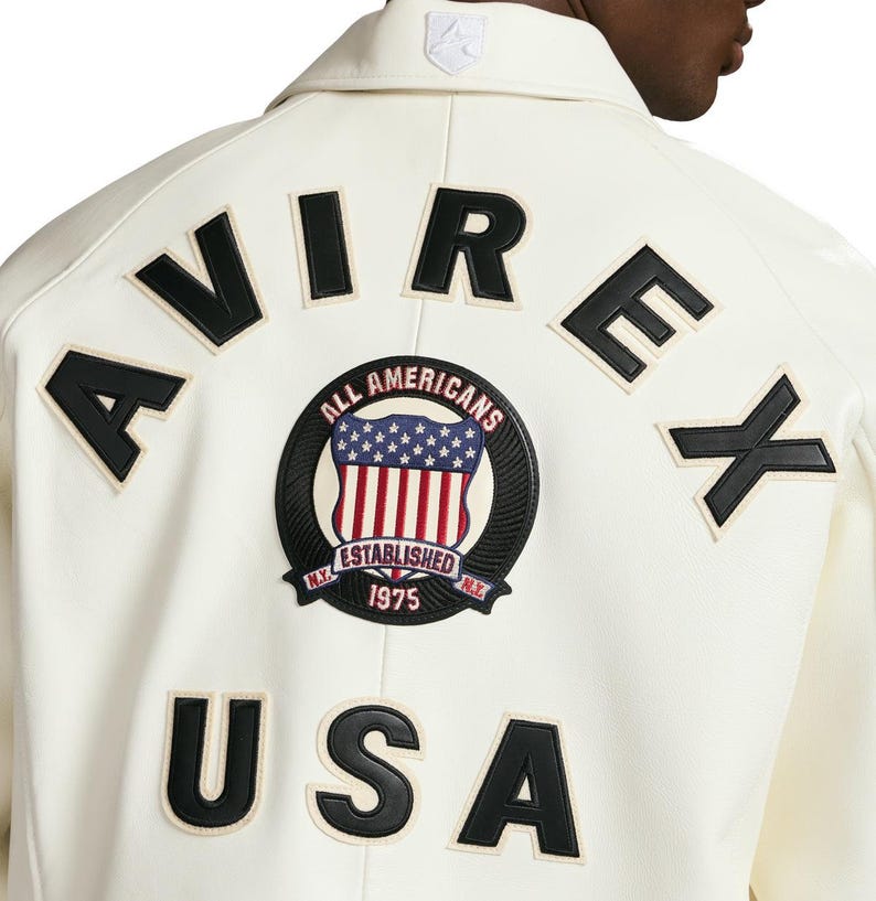 Op de afbeelding: Een gebroken witte jas met zwarte leren letters die AVIREX en USA op de achterkant vormen. Een ronde geborduurde patch in het midden toont een schild met sterren en strepen, en de tekst ALL AMERICANS, ESTABLISHED en 1975.