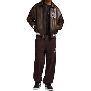 May include: A brown leather bomber jacket with a collar, white striped cuffs, and a patch featuring a flag design on the chest. The jacket is worn over a dark t-shirt and dark brown sweatpants with a white graphic, paired with black and white trainers.