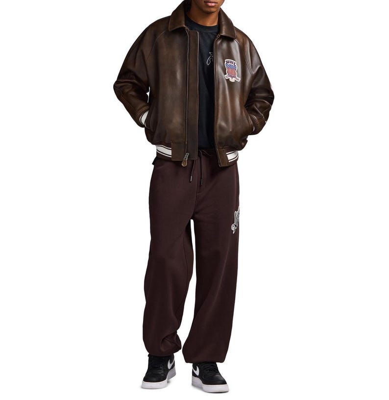 May include: A brown leather bomber jacket with a collar, white striped cuffs, and a patch featuring a flag design on the chest. The jacket is worn over a dark t-shirt and dark brown sweatpants with a white graphic, paired with black and white trainers.