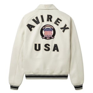 Op de afbeelding: Een gebroken witte jas met zwarte letters AVIREX en USA op de achterkant. Een ronde patch in het midden toont de tekst ALL AMERICANS ESTABLISHED 1975 met een rood, wit en blauw vlagontwerp. De jas heeft boorden met strepen.
