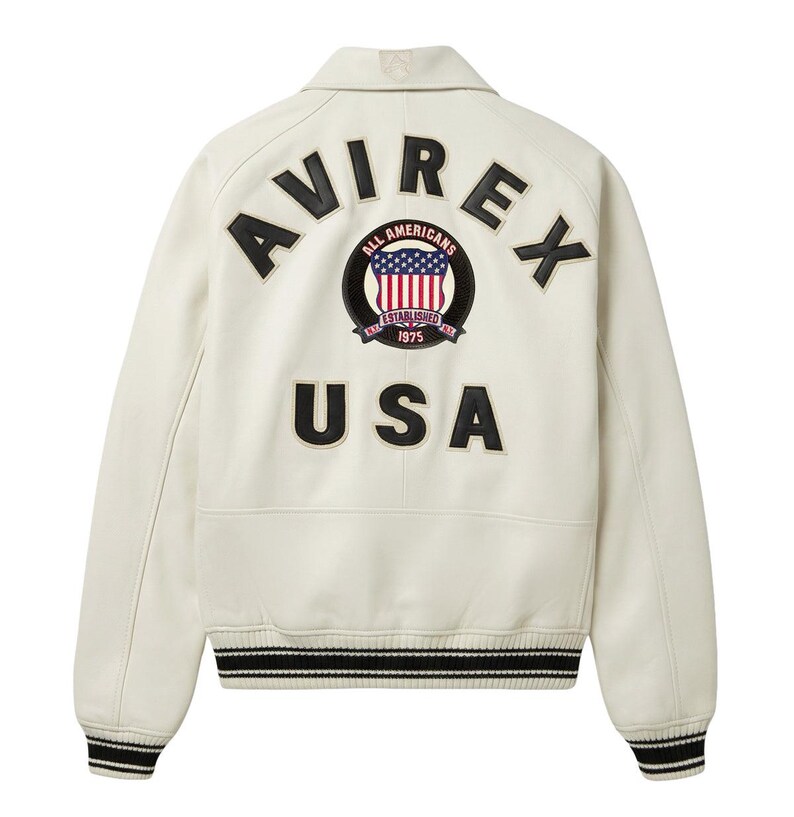 Op de afbeelding: Een gebroken witte jas met zwarte letters AVIREX en USA op de achterkant. Een ronde patch in het midden toont de tekst ALL AMERICANS ESTABLISHED 1975 met een rood, wit en blauw vlagontwerp. De jas heeft boorden met strepen.