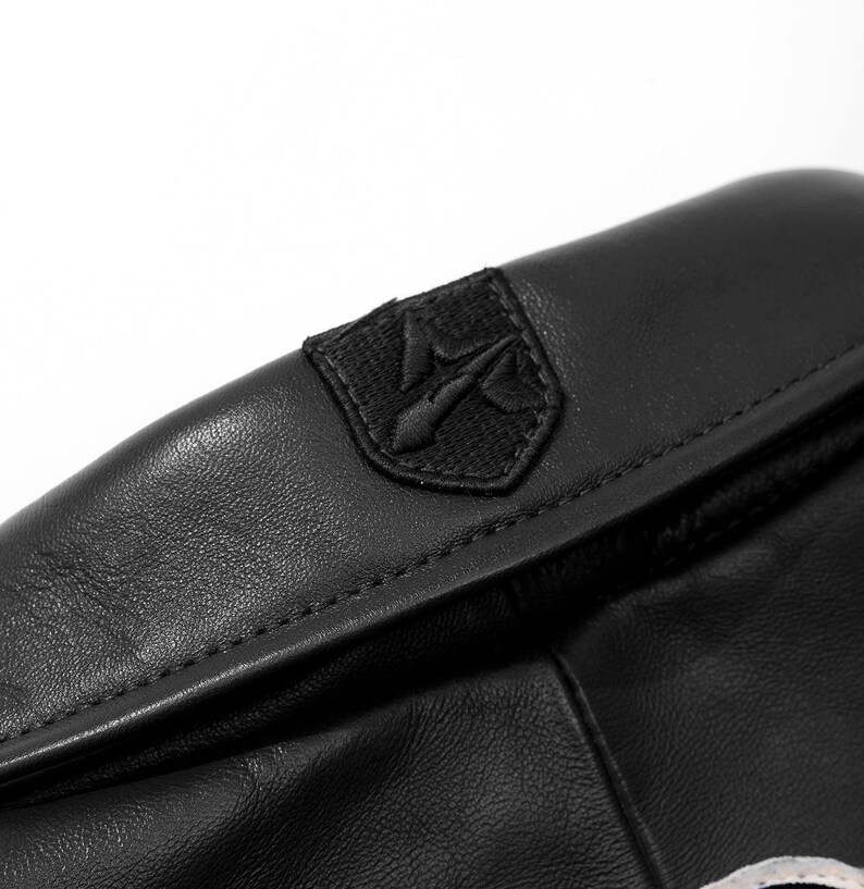 May include: A black leather item featuring a smooth, matte texture and a stitched seam along the top edge. Centered on the upper portion is a black embroidered patch in the shape of a shield, displaying a stylised trident symbol in black thread.