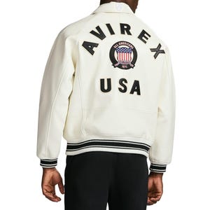 Op de afbeelding: Een off-white varsity jack met zwarte leren letters die AVIREX vormen in een boog over de bovenrug. Onder de tekst bevindt zich een ronde badge met een rood, wit en blauw vlagontwerp, gevolgd door de zwarte leren letters U S A.