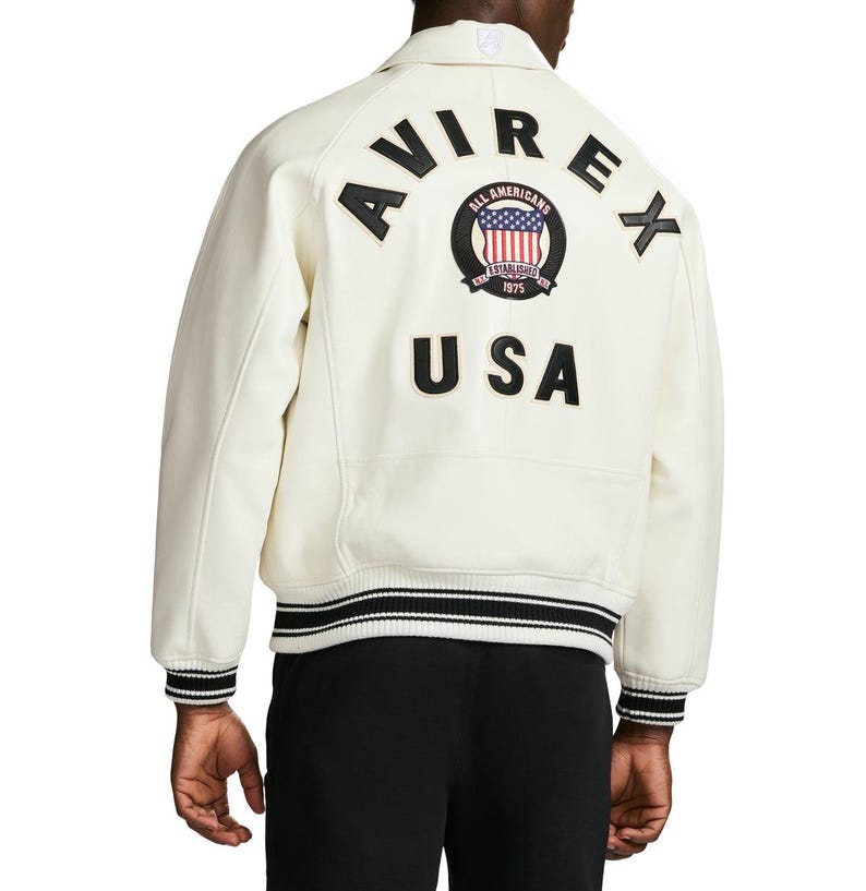Op de afbeelding: Een off-white varsity jack met zwarte leren letters die AVIREX vormen in een boog over de bovenrug. Onder de tekst bevindt zich een ronde badge met een rood, wit en blauw vlagontwerp, gevolgd door de zwarte leren letters U S A.