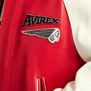May include: A red and white leather varsity jacket featuring the text AVIREX in black embroidery above a black and white embroidered patch of a winged profile of a person. The jacket has a smooth texture and a vertical pocket with white trim.