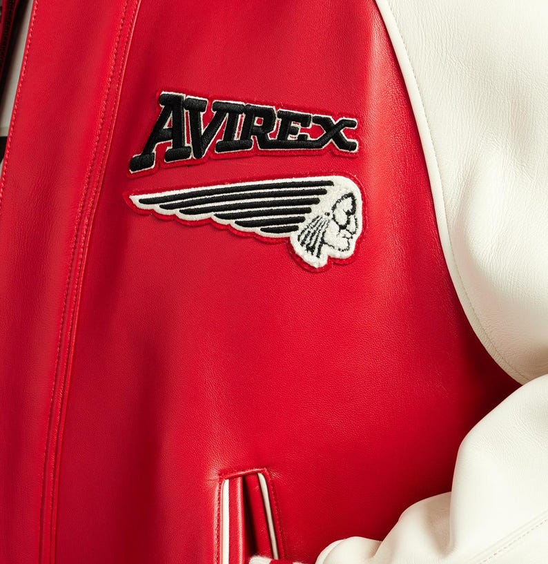 May include: A red and white leather varsity jacket featuring the text AVIREX in black embroidery above a black and white embroidered patch of a winged profile of a person. The jacket has a smooth texture and a vertical pocket with white trim.