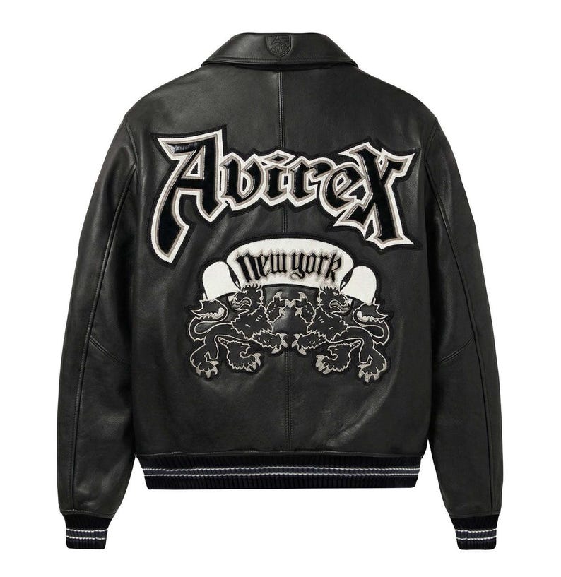 May include: A black leather jacket featuring a large embroidered Avirex logo across the back in white and black. Below the logo is a banner with the text New York and an emblem of two lions facing each other. The jacket has a ribbed collar and cuffs.