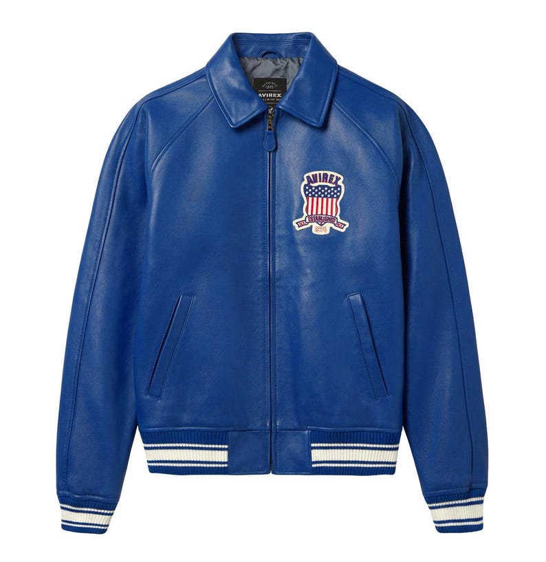 May include: A bright blue leather jacket featuring a classic collar, a full-length front zip, and two side pockets. On the left chest, there is an embroidered patch displaying the text AVIREX above a shield with a flag design and the word ESTABLISHED.