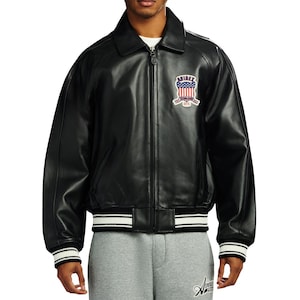 May include: A black leather bomber jacket featuring a classic collar, a full-length front zip, and ribbed cuffs and hem with white stripes. A patch on the left chest displays the text AVIREX with a red, white, and blue American flag shield.