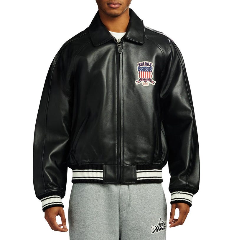 May include: A black leather bomber jacket featuring a classic collar, a full-length front zip, and ribbed cuffs and hem with white stripes. A patch on the left chest displays the text AVIREX with a red, white, and blue American flag shield.