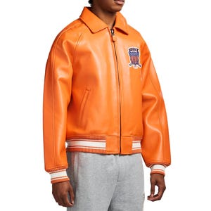 May include: A bright orange leather jacket featuring a front zipper closure, a classic collar, and ribbed cuffs and hem with white stripes. An embroidered patch on the chest displays the text AVIREX above a shield with a red, white, and blue flag design.