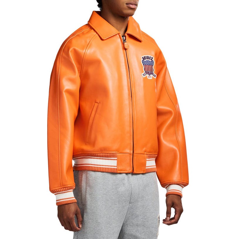 May include: A bright orange leather jacket featuring a front zipper closure, a classic collar, and ribbed cuffs and hem with white stripes. An embroidered patch on the chest displays the text AVIREX above a shield with a red, white, and blue flag design.