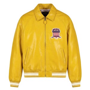 May include: A mustard yellow leather jacket featuring a front zipper closure, a classic collar, and ribbed cuffs and hem with white stripes. On the left chest, there is a patch with the text AVIREX ESTABLISHED 1975 and a US flag shield design.