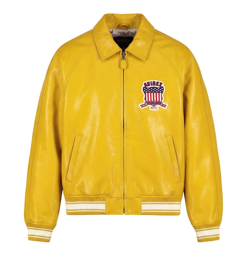 May include: A mustard yellow leather jacket featuring a front zipper closure, a classic collar, and ribbed cuffs and hem with white stripes. On the left chest, there is a patch with the text AVIREX ESTABLISHED 1975 and a US flag shield design.