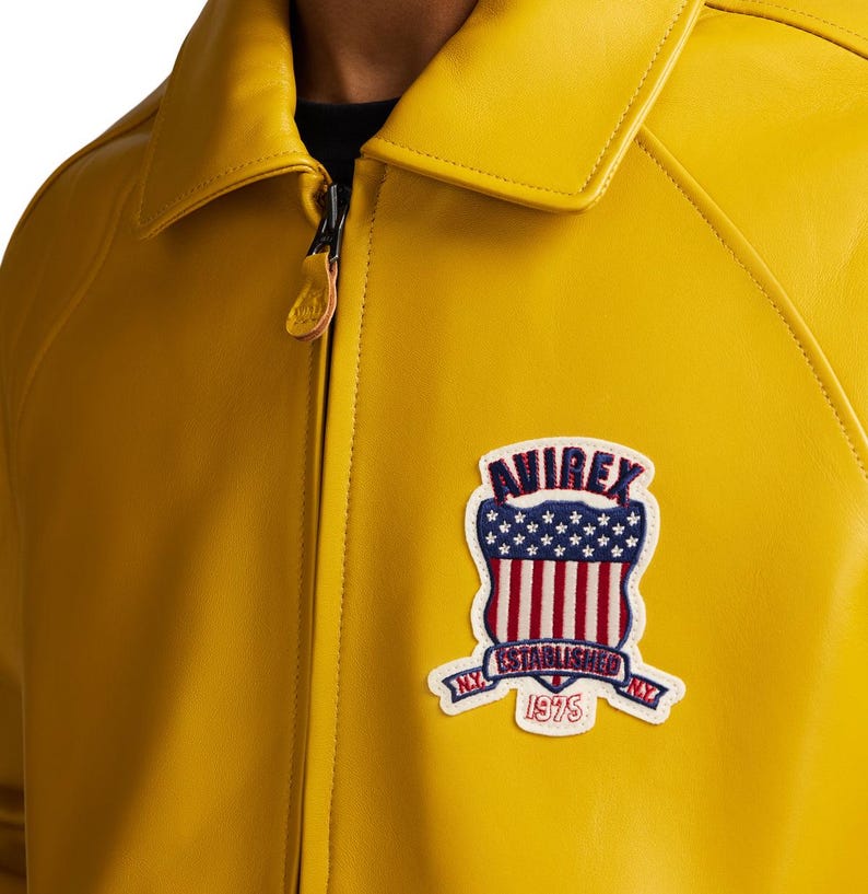 May include: A bright yellow leather jacket with a full-length front zipper and a pointed collar. On the left chest, there is a shield-shaped patch featuring the text AVIREX, ESTABLISHED 1975, and N.Y., along with a red, white, and blue flag design.