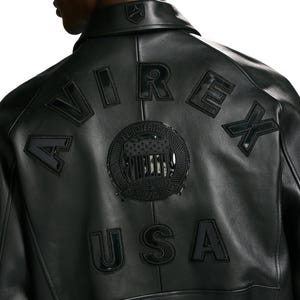 Puede incluir: Una chaqueta de cuero negra que presenta la palabra AVIREX arqueada en la parte superior de la espalda en letras negras grandes. Debajo, un emblema circular con un dise&ntilde;o de bandera estrellada est&aacute; centrado, con las letras USA debajo.