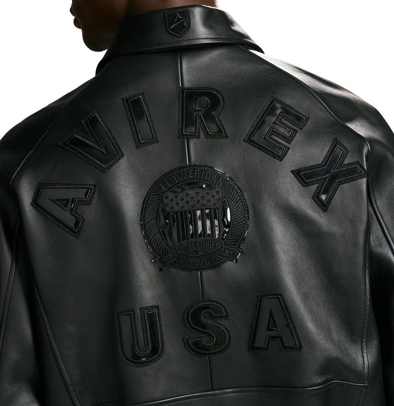 Puede incluir: Una chaqueta de cuero negra que presenta la palabra AVIREX arqueada en la parte superior de la espalda en letras negras grandes. Debajo, un emblema circular con un dise&ntilde;o de bandera estrellada est&aacute; centrado, con las letras USA debajo.