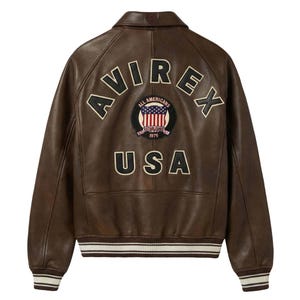 May include: A brown leather jacket featuring the word AVIREX arched across the upper back in black letters with cream trim. Below is a circular patch with an American flag and the text ALL AMERICANS ESTABLISHED 1975. USA is printed below.