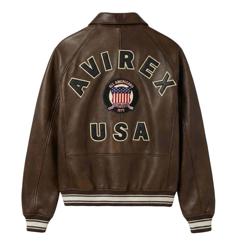 May include: A brown leather jacket featuring the word AVIREX arched across the upper back in black letters with cream trim. Below is a circular patch with an American flag and the text ALL AMERICANS ESTABLISHED 1975. USA is printed below.