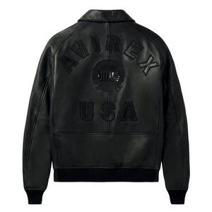Puede incluir: Una chaqueta de cuero negra con cuello vuelto y pu&ntilde;os y dobladillo acanalados. La parte posterior muestra letras en relieve que dicen AVIREX USA, junto con un emblema circular centrado entre las palabras, todo en un acabado negro a juego.