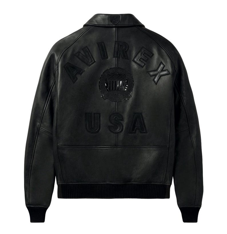 Puede incluir: Una chaqueta de cuero negra con cuello vuelto y pu&ntilde;os y dobladillo acanalados. La parte posterior muestra letras en relieve que dicen AVIREX USA, junto con un emblema circular centrado entre las palabras, todo en un acabado negro a juego.