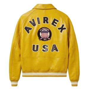 May include: A mustard yellow leather jacket featuring a ribbed collar, cuffs, and hem with white stripes. The back displays the black text AVIREX and USA, with a circular patch in the center depicting an American flag and the text ALL AMERICANS.
