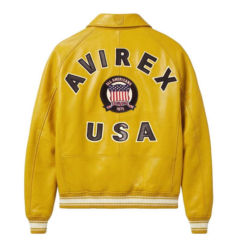 May include: A mustard yellow leather jacket featuring a ribbed collar, cuffs, and hem with white stripes. The back displays the black text AVIREX and USA, with a circular patch in the center depicting an American flag and the text ALL AMERICANS.
