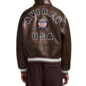 May include: A brown leather bomber jacket featuring the text AVIREX in an arch, a circular patch with an American flag and the text ALL AMERICANS ESTABLISHED 1975, and the text USA below it. The jacket has a ribbed collar and cuffs with white stripes.