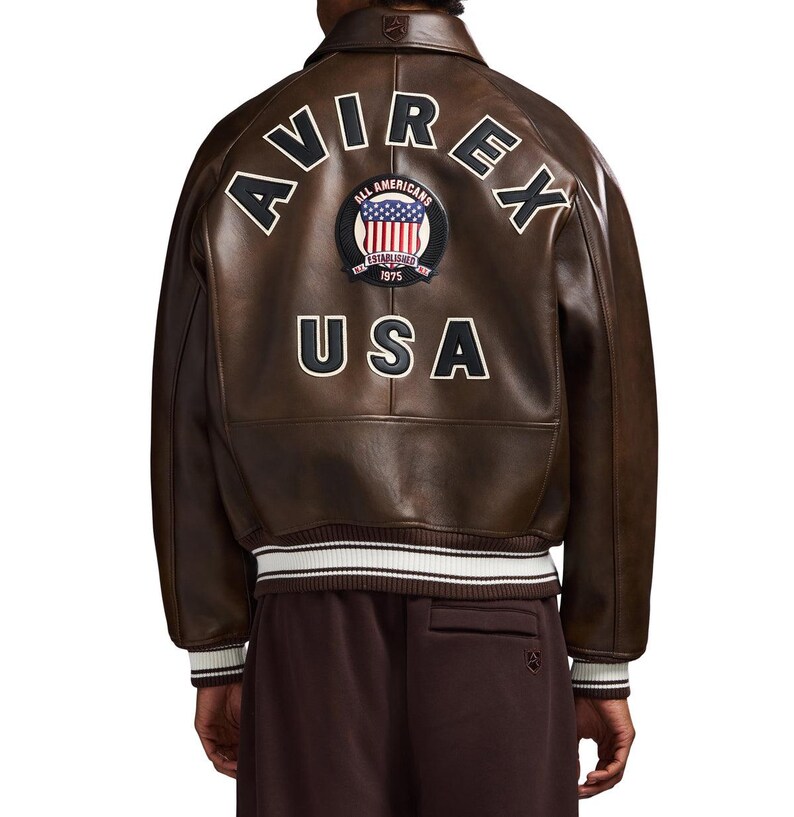 May include: A brown leather bomber jacket featuring the text AVIREX in an arch, a circular patch with an American flag and the text ALL AMERICANS ESTABLISHED 1975, and the text USA below it. The jacket has a ribbed collar and cuffs with white stripes.
