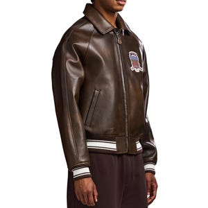 May include: A dark brown leather bomber jacket featuring a classic collar, a front zip closure with a branded pull, and ribbed cuffs and hem with white stripes. A patch on the chest displays the text AVIREX with a red, white, and blue shield.