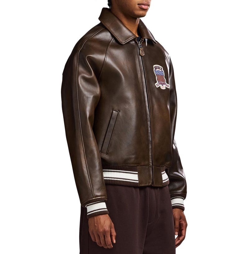 May include: A dark brown leather bomber jacket featuring a classic collar, a front zip closure with a branded pull, and ribbed cuffs and hem with white stripes. A patch on the chest displays the text AVIREX with a red, white, and blue shield.