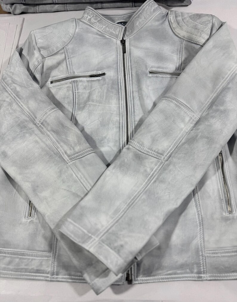K&ouml;nnte beinhalten: Hellgraue Lederjacke mit Rei&szlig;verschluss vorne und mehreren Rei&szlig;verschlusstaschen. Die Jacke hat gesteppte Schulterdetails und einen Stehkragen. Die &Auml;rmel sind &uuml;ber der Vorderseite gefaltet.