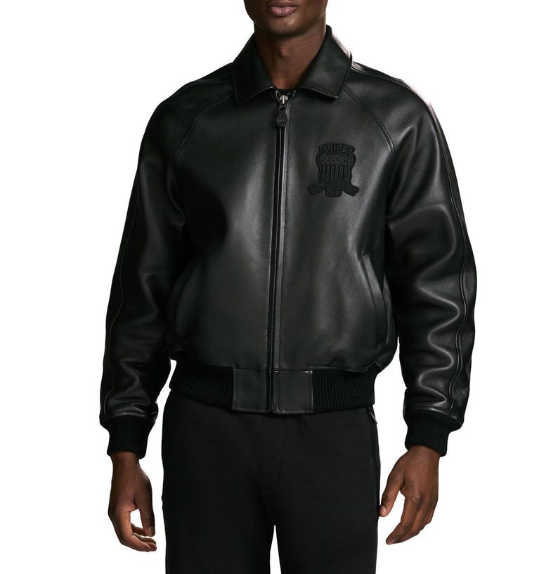 Puede incluir: Una chaqueta bomber de cuero negro con cuello cl&aacute;sico, cierre de cremallera frontal completo y pu&ntilde;os y dobladillo de punto acanalado. El pecho presenta un emblema bordado en negro tonal con un dise&ntilde;o de escudo y rayas horizontales.