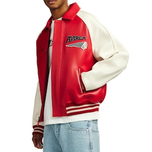 May include: A red and white varsity jacket featuring the text AVIREX with a winged emblem on the chest. The jacket has a classic collar, long sleeves, and a ribbed hem with white stripes. It is worn over a white t-shirt and light blue denim jeans.