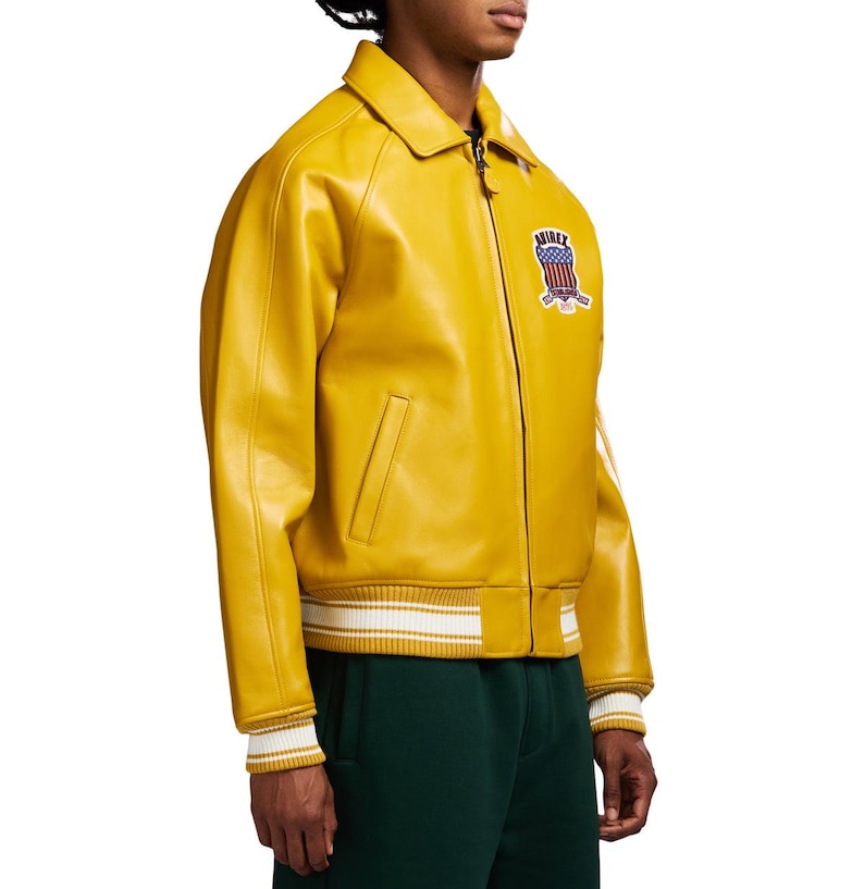 May include: A mustard yellow leather jacket featuring a classic collar, a front zipper closure, and two side pockets. The chest displays a patch with the text AVIREX and a graphic of the American flag. The cuffs and hem have white striped ribbing.