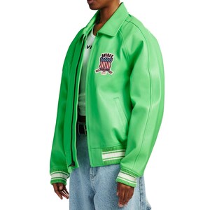 K&ouml;nnte beinhalten: Eine leuchtend lindgr&uuml;ne Bomberjacke mit klassischem Kragen, Frontrei&szlig;verschluss und gerippten B&uuml;ndchen mit wei&szlig;en Streifen. Ein gestickter Aufn&auml;her auf der Brust zeigt den Schriftzug AVIREX &uuml;ber einem Wappen mit rot-wei&szlig;-blauem Flaggendesign.