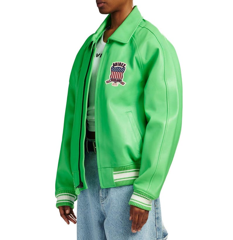 K&ouml;nnte beinhalten: Eine leuchtend lindgr&uuml;ne Bomberjacke mit klassischem Kragen, Frontrei&szlig;verschluss und gerippten B&uuml;ndchen mit wei&szlig;en Streifen. Ein gestickter Aufn&auml;her auf der Brust zeigt den Schriftzug AVIREX &uuml;ber einem Wappen mit rot-wei&szlig;-blauem Flaggendesign.