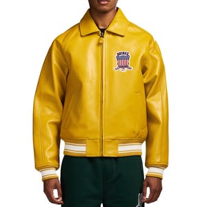 May include: A mustard yellow leather bomber jacket with a pointed collar, a full-length front zipper, and ribbed cuffs and hem featuring white stripes. A patch on the left chest displays the text AVIREX above a shield with a red, white, and blue flag.